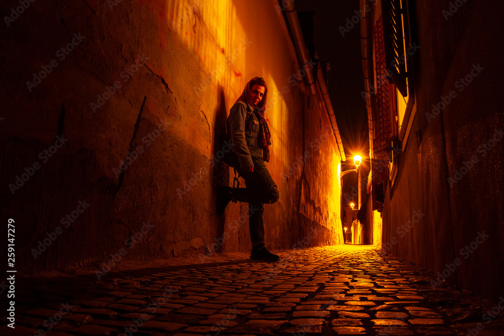 Moody portrait of a young woman on a narrow urban passage, at night, next to open window shutters, in Sibiu (Hermannstadt), with scarce street lights reflecting yellow/orange rays on the cobbles.