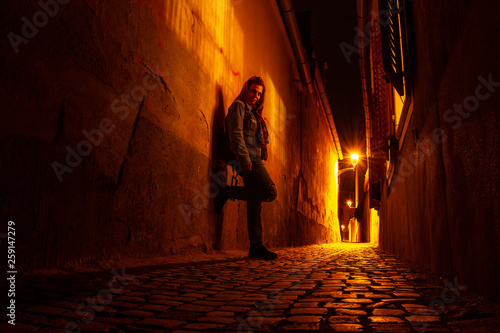 Obraz na płótnie Moody portrait of a young woman on a narrow urban passage, at night, next to open window shutters, in Sibiu (Hermannstadt), with scarce street lights reflecting yellow/orange rays on the cobbles. w salonie