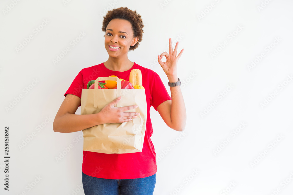 Young african american woman holding paper bag full of fresh groceries doing ok sign with fingers, excellent symbol Young african american woman holding paper bag full of fresh groceries doing ok sign with fingers, excellent symbol