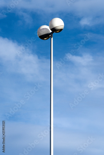 High tall lamp post streetlight with futuristic two spherical round light fixture on the upper end with blue sky and white clouds in the background - concept lantern illumination modern design future 