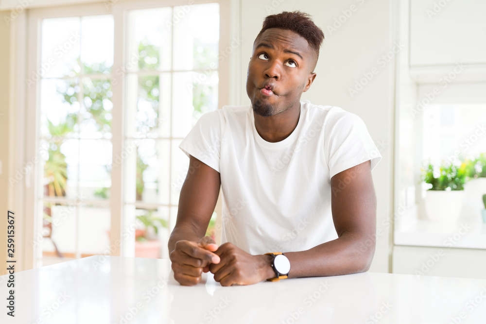 Handsome african american man on white table at home making fish face with lips, crazy and comical gesture. Funny expression. Handsome african american man on white table at home making fish face with lips, crazy and comical gesture. Funny expression.