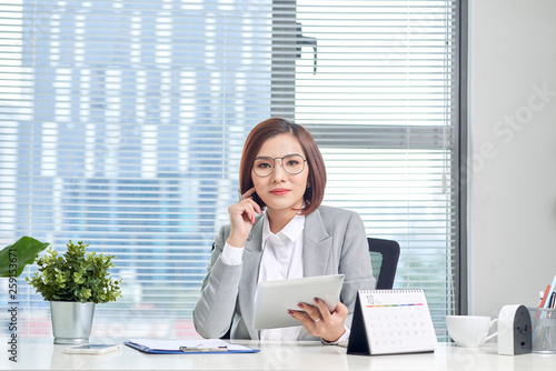 Portrait of beautiful young Asian business woman sitting and using a tablet behind the table in workplace