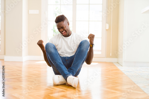 Handsome african american man sitting on the floor at home very happy and excited doing winner gesture with arms raised, smiling and screaming for success. Celebration concept. © Krakenimages.com