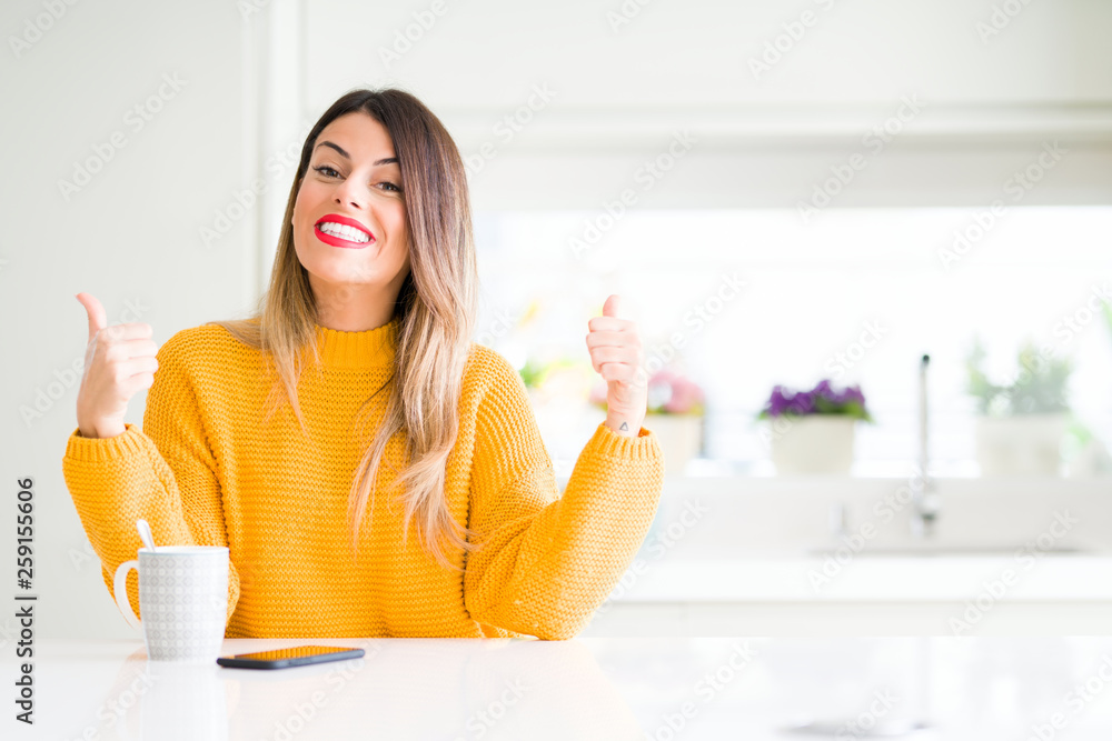 Young beautiful woman drinking a cup of coffee at home success sign doing positive gesture with hand, thumbs up smiling and happy. Looking at the camera with cheerful expression, winner gesture. Young beautiful woman drinking a cup of coffee at home success sign doing positive gesture with hand, thumbs up smiling and happy. Looking at the camera with cheerful expression, winner gesture.