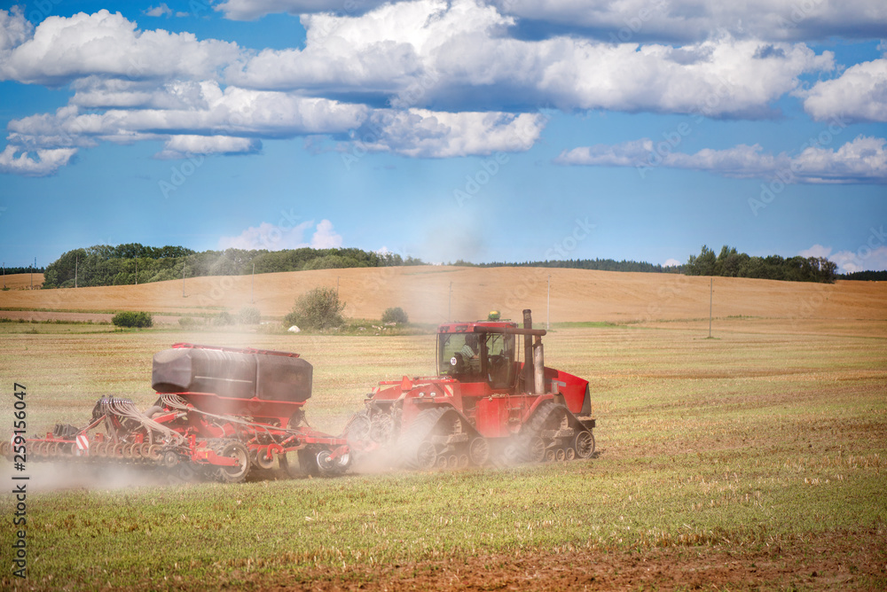 Fototapeta premium Agricultural background with red tractor pulling plow, throwing dust in air. Combine harvester at wheat field. Heavy machinery during cultivation, working on fields. Dramatic sky, rain, storm clouds 