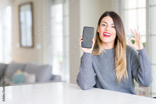 Young beautiful woman showing smartphone screen at home doing ok sign with fingers, excellent symbol