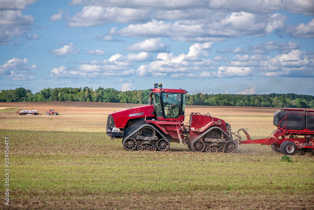 Obraz premium Agricultural background with red tractor pulling plow, throwing dust in air. Combine harvester at wheat field. Heavy machinery during cultivation, working on fields. Dramatic sky, rain, storm clouds