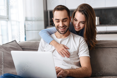 Image of joyful couple using laptop together while sitting on sofa in living room at home