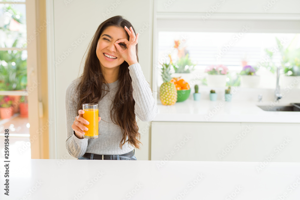 Young woman drinking a glass of fresh orange juice with happy face smiling doing ok sign with hand on eye looking through fingers Young woman drinking a glass of fresh orange juice with happy face smiling doing ok sign with hand on eye looking through fingers