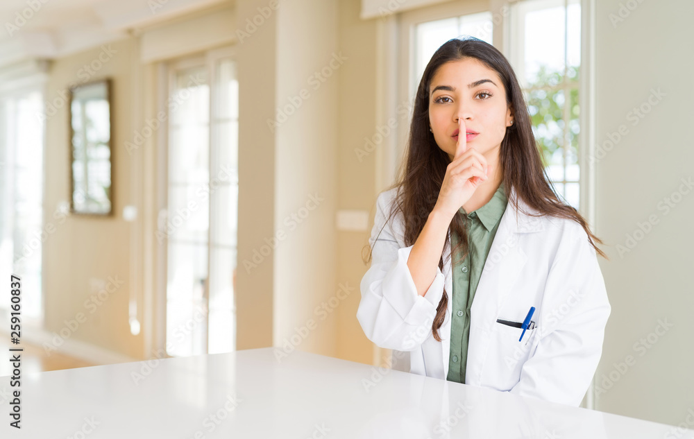 Young woman wearing medical coat at the clinic as therapist or doctor asking to be quiet with finger on lips. Silence and secret concept.
