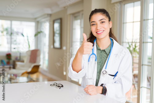 Young doctor woman wearing medical coat at the clinic doing happy thumbs up gesture with hand. Approving expression looking at the camera with showing success.