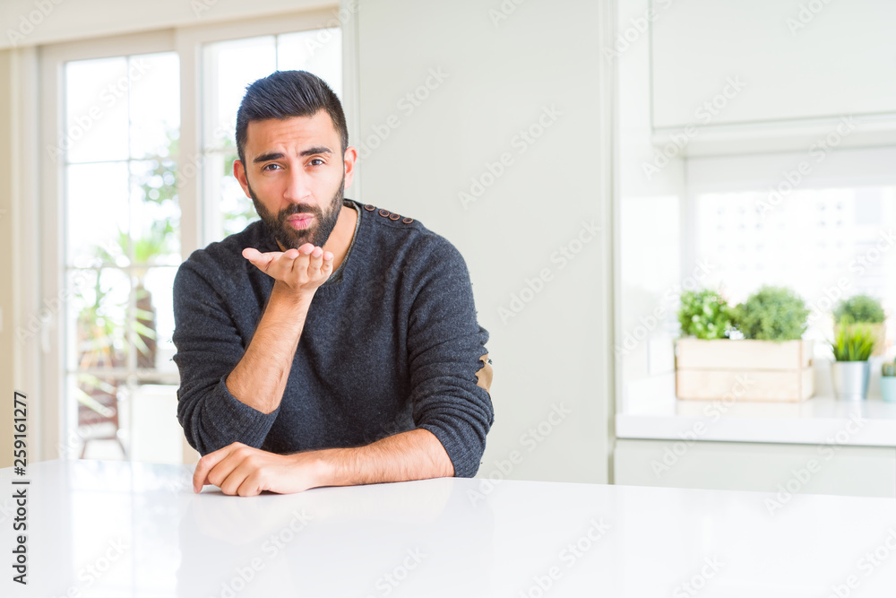 Handsome hispanic man wearing casual sweater at home looking at the camera blowing a kiss with hand on air being lovely and sexy. Love expression.