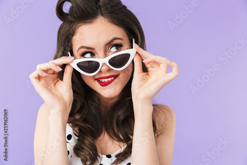 Portrait of trendy pin-up woman 20s in vintage polka dot dress and retro sunglasses smiling at camera