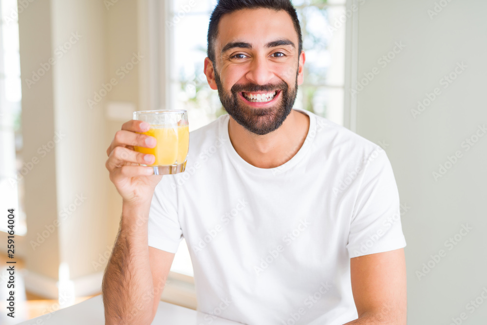 Handsome hispanic man drinking healthy orange juice with a happy face standing and smiling with a confident smile showing teeth Handsome hispanic man drinking healthy orange juice with a happy face standing and smiling with a confident smile showing teeth
