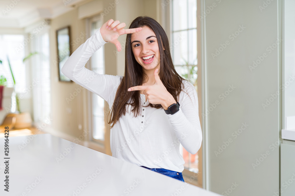 Beautiful young woman sitting on white table at home smiling making frame with hands and fingers with happy face. Creativity and photography concept. Beautiful young woman sitting on white table at home smiling making frame with hands and fingers with happy face. Creativity and photography concept.