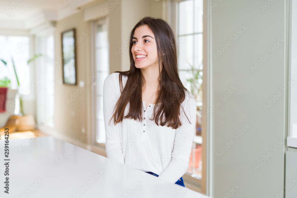 Beautiful young woman sitting on white table at home looking away to side with smile on face, natural expression. Laughing confident.