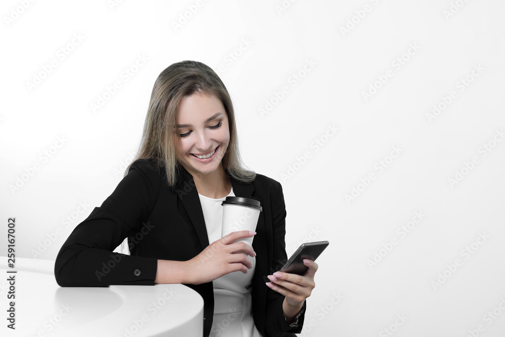 A young beautiful woman in a black suit and white blouse is sitting at the office table. Drinks a hot drink of coffee from a white paper cup. Writes a message on a smartphone, communicates and smiles. A young beautiful woman in a black suit and white blouse is sitting at the office table. Drinks a hot drink of coffee from a white paper cup. Writes a message on a smartphone, communicates and smiles.