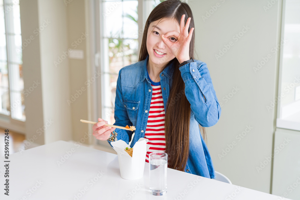 Beautiful Asian woman eating asian rice in delivery box with happy face smiling doing ok sign with hand on eye looking through fingers Beautiful Asian woman eating asian rice in delivery box with happy face smiling doing ok sign with hand on eye looking through fingers