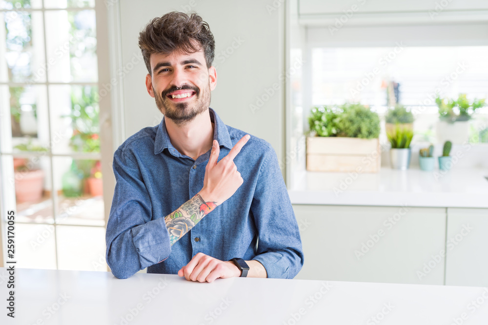 Young man wearing casual shirt sitting on white table cheerful with a smile of face pointing with hand and finger up to the side with happy and natural expression on face Young man wearing casual shirt sitting on white table cheerful with a smile of face pointing with hand and finger up to the side with happy and natural expression on face