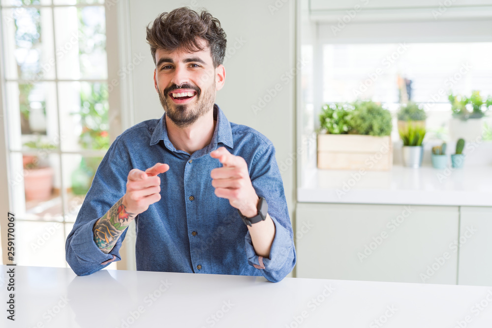 Young man wearing casual shirt sitting on white table pointing fingers to camera with happy and funny face. Good energy and vibes. Young man wearing casual shirt sitting on white table pointing fingers to camera with happy and funny face. Good energy and vibes.