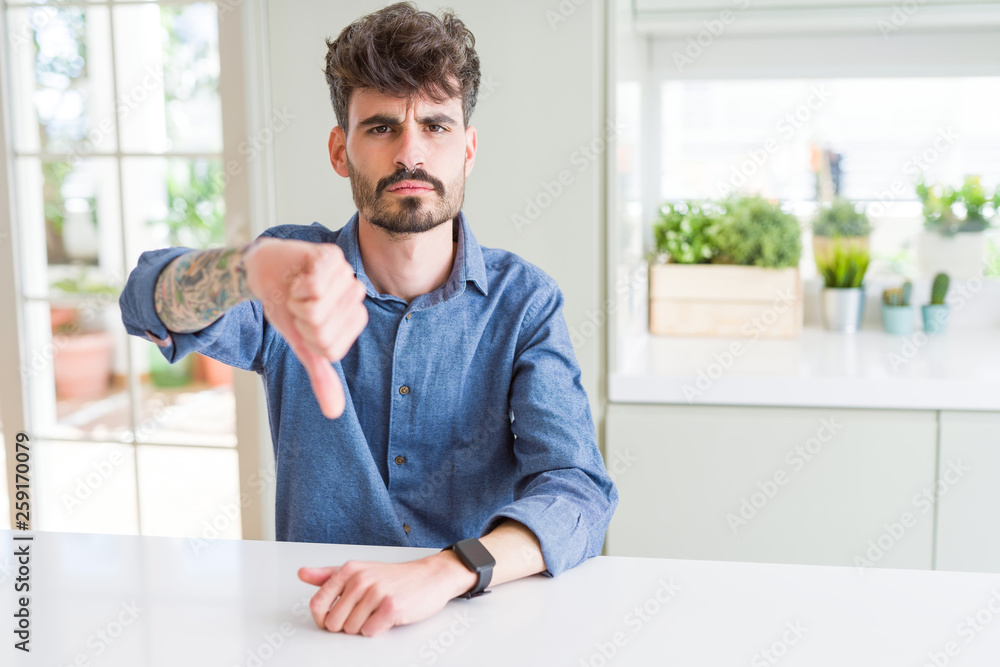 Fototapeta premium Young man wearing casual shirt sitting on white table looking unhappy and angry showing rejection and negative with thumbs down gesture. Bad expression.