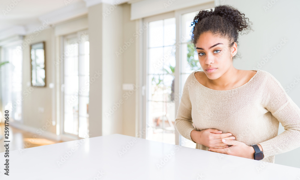 Obraz premium Beautiful young african american woman with afro hair sitting on table at home with hand on stomach because nausea, painful disease feeling unwell. Ache concept.