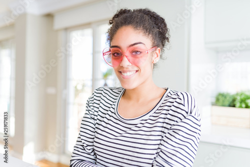 Beautiful young african american woman with afro hair wearing crazy pink heart glasses with a happy and cool smile on face. Lucky person.