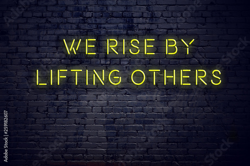Positive inspiring quote on neon sign against brick wall we rise by lifting others