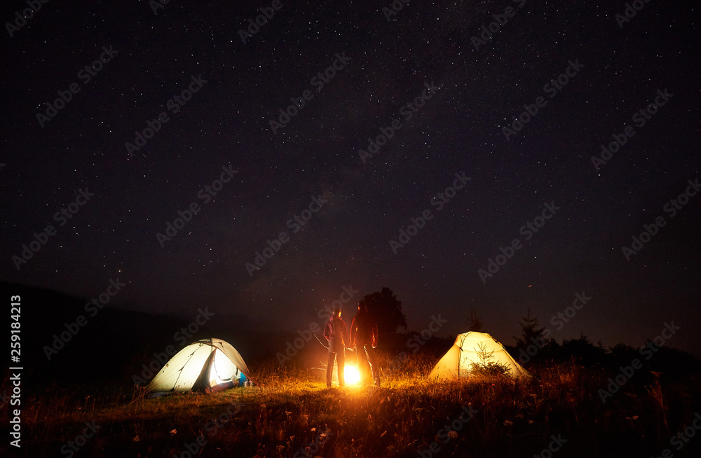 Fototapeta premium Night camping picture. Bright bonfire burning between two silhouettes of boy and girl standing in front of illulminated tents, holding hands under beautiful dark starry sky on distant hills background