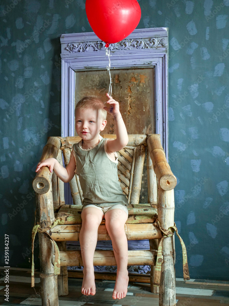 Fototapeta premium Funny boy in blue clothes and glasses is playing with red balloon against gray wall against background of ancient mirror. child is happy at home