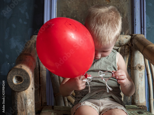 Funny boy in blue clothes and glasses is playing with red balloon against gray wall against background of ancient mirror. child is happy at home