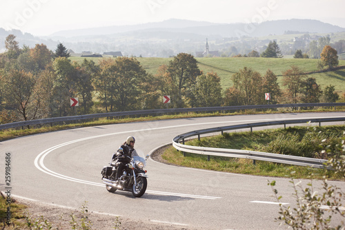Bearded motorcyclist in helmet, sunglasses and black leather clothing riding bike along sharp turn of empty road on bright summer day on misty background of rural landscape and distant green hills. © anatoliy_gleb