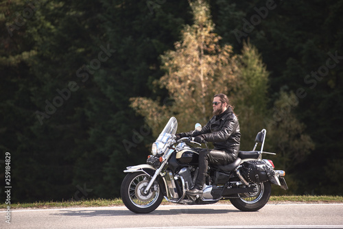Side view of bearded long-haired motorcyclist in sunglasses and black leather clothing riding cruiser motorbike along narrow asphalt path on sunny autumn day on background of forest.