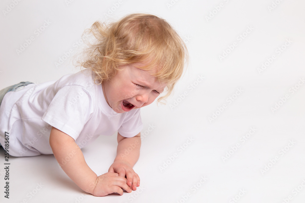 A little curly crying child in a clean white T-shirt is lying on the floor and crying. A little curly crying child in a clean white T-shirt is lying on the floor and crying.