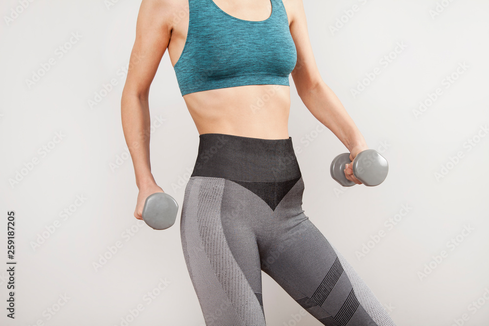 Fototapeta premium Sporty woman does the exercises with dumbbells on gray background.