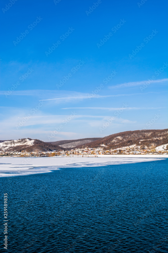 Small European village on the side of a frozen lake with dark blue water and clear sky. Winter holidays and vacation, Christmas and New Year recreation, beautiful landscape.