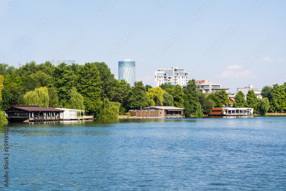 Herastrau lake and park in summer with Bucharest skyline formed of office towers in the background. Urban leisure and recreation area with blue water and green trees. 
