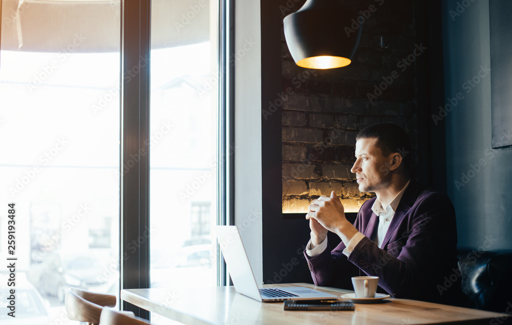 Young handsome man sitting in office with cup of coffee and working on project connected with modern cyber technologies. Businessman with notebook trying to keep deadline in digital marketing sphere. Young handsome man sitting in office with cup of coffee and working on project connected with modern cyber technologies. Businessman with notebook trying to keep deadline in digital marketing sphere.