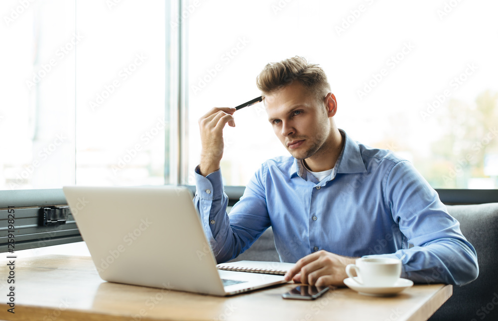 Young handsome man sitting in office with cup of coffee and working on project connected with modern cyber technologies. Businessman with notebook trying to keep deadline in digital marketing sphere. Young handsome man sitting in office with cup of coffee and working on project connected with modern cyber technologies. Businessman with notebook trying to keep deadline in digital marketing sphere.