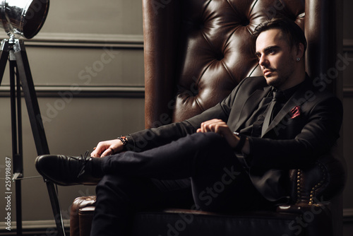 Young handsome man posing for a fashion shoot in a studio. Fashion as a lifestyle. Man wearing a beard. Model sitting on a sofa in tuxedo. Successful fashionable businessman. Business look advertising