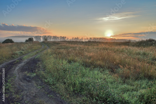 morning, rising sun over the field and dirt road, with trees and country buildings