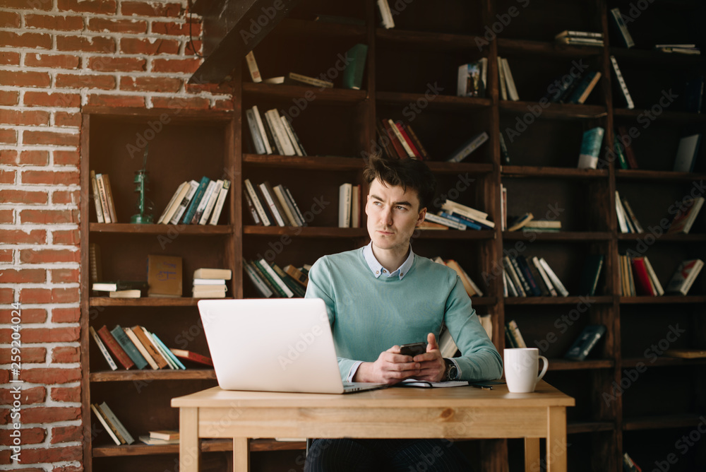 Young handsome man sitting in office with cup of coffee and working on project connected with modern cyber technologies. Businessman with notebook trying to keep deadline in digital marketing sphere. Young handsome man sitting in office with cup of coffee and working on project connected with modern cyber technologies. Businessman with notebook trying to keep deadline in digital marketing sphere.