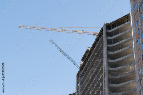 high-rise construction crane with a long arrow of yellow color against the blue sky over a new multi-storey building of concrete and brick under construction © Тарас Квакуш