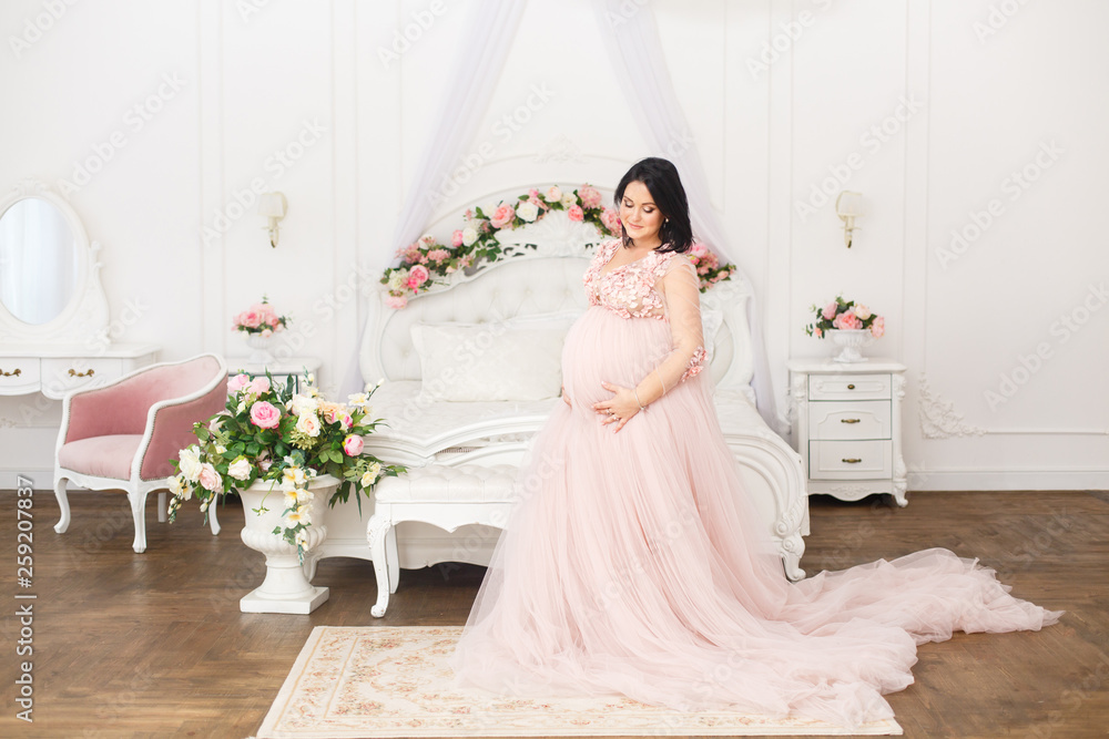 Young pregnant woman in a light classic interior. Caucasian woman with black hairs in long romantic dress near the large window. Concept of new life. Waiting of baby. Pretty pregnant woman in a room Young pregnant woman in a light classic interior. Caucasian woman with black hairs in long romantic dress near the large window. Concept of new life. Waiting of baby. Pretty pregnant woman in a room