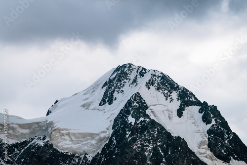 Wonderful glacier close-up. Snowy dark mountain top in cloudy sky. Rocky ridge with snow in overcast weather. Amazing mountain range in mist. Atmospheric minimalist landscape of majestic nature.