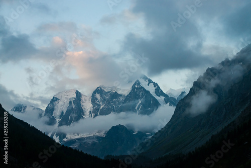 Low cloud before huge glacier. Giant snowy rocky mountains under cloudy sky. Thick mist in mountains above forest at early morning. Impenetrable fog. Dark atmospheric landscape. Tranquil atmosphere.
