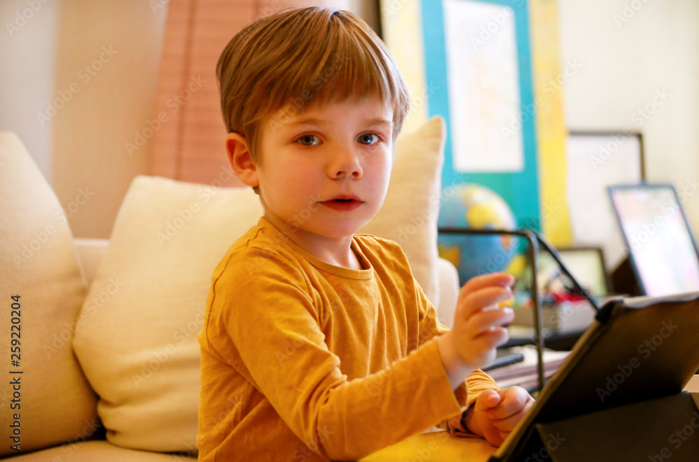 Child using tablet pc on bed at home. Cute boy on sofa is watching cartoon, playing games and learning from laptop. Education, fun, leisure, happiness, modern computer technology and communication. Child using tablet pc on bed at home. Cute boy on sofa is watching cartoon, playing games and learning from laptop. Education, fun, leisure, happiness, modern computer technology and communication.