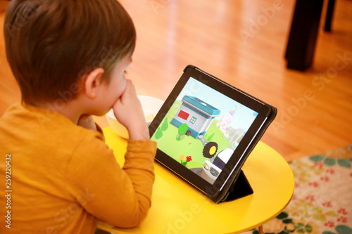 Child using tablet pc on bed at home. Cute boy on sofa is watching cartoon, playing games and learning from laptop. Education, fun, leisure, happiness, modern computer technology and communication.