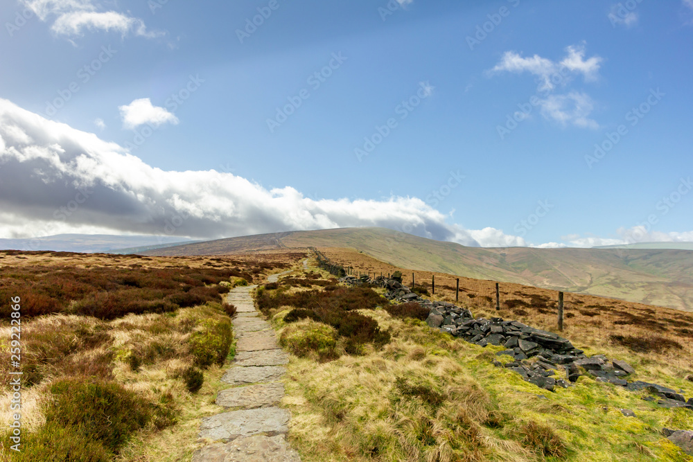 A view of a stony path trail along heater in the hill under a majestic blue sky and white clouds