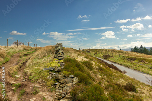 A view of a muddy trail path along a stone wall and green vegetation under a majestic blue sky and white clouds © Dolwolfian